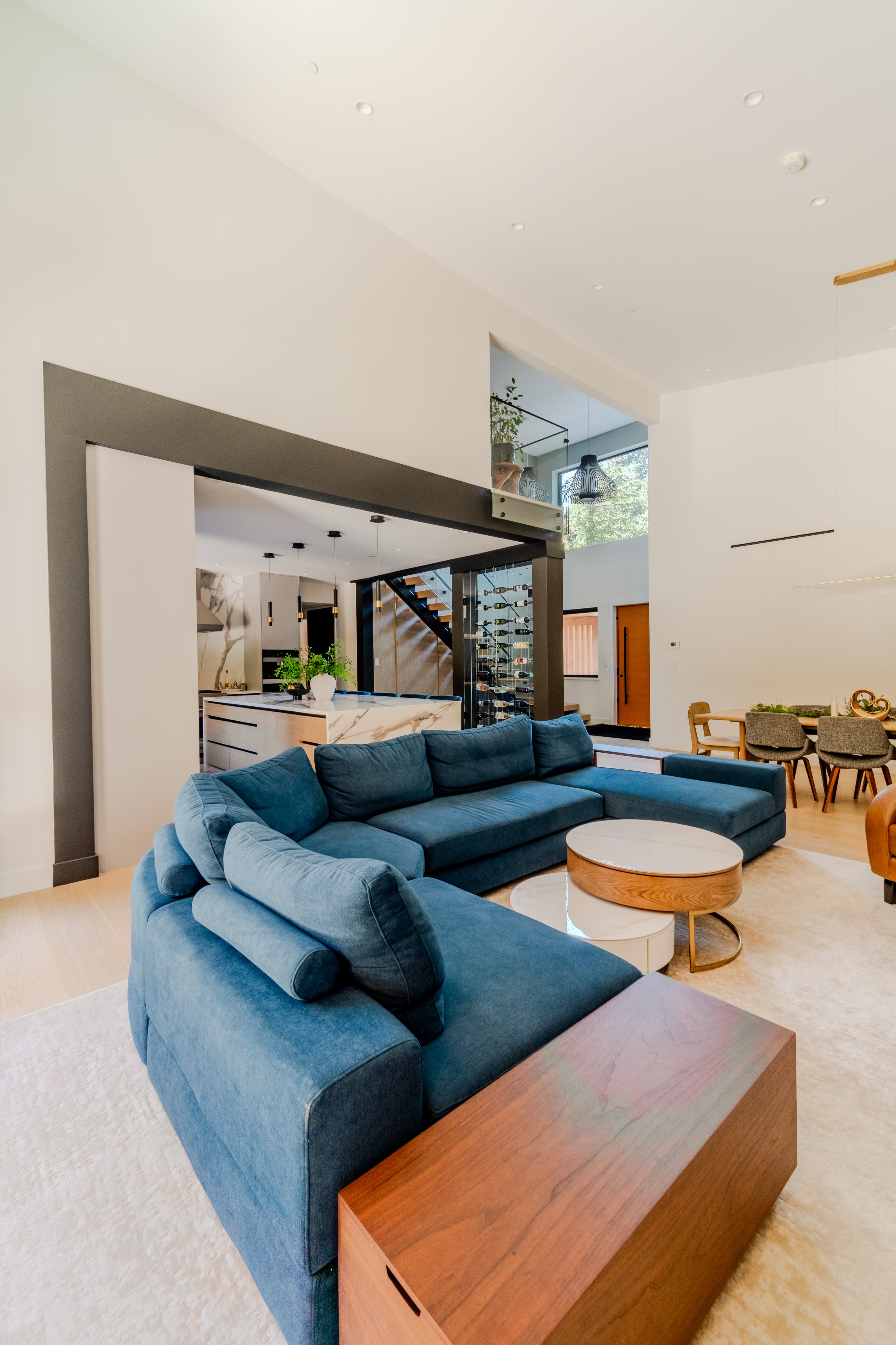 Open-plan living room real estate photography Vancouver — oversized blue velvet sectional, round gold coffee table, double-height ceilings, open-tread staircase, and open-plan kitchen beyond