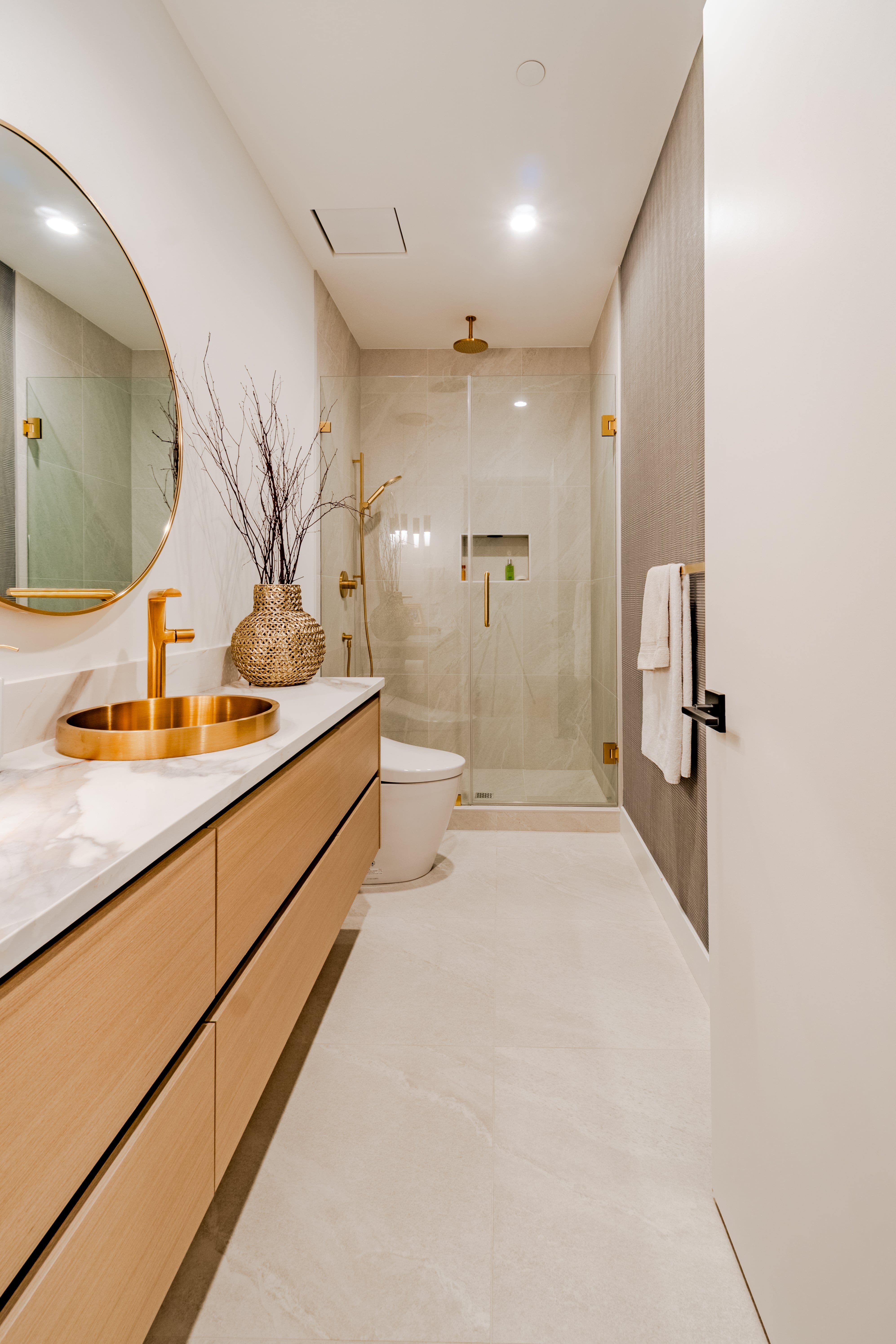 Modern ensuite real estate photography Vancouver — floating light wood vanity with white marble countertop, round brass mirror, brushed gold vessel sink and faucet, glass shower with gold fixtures, and large-format tile