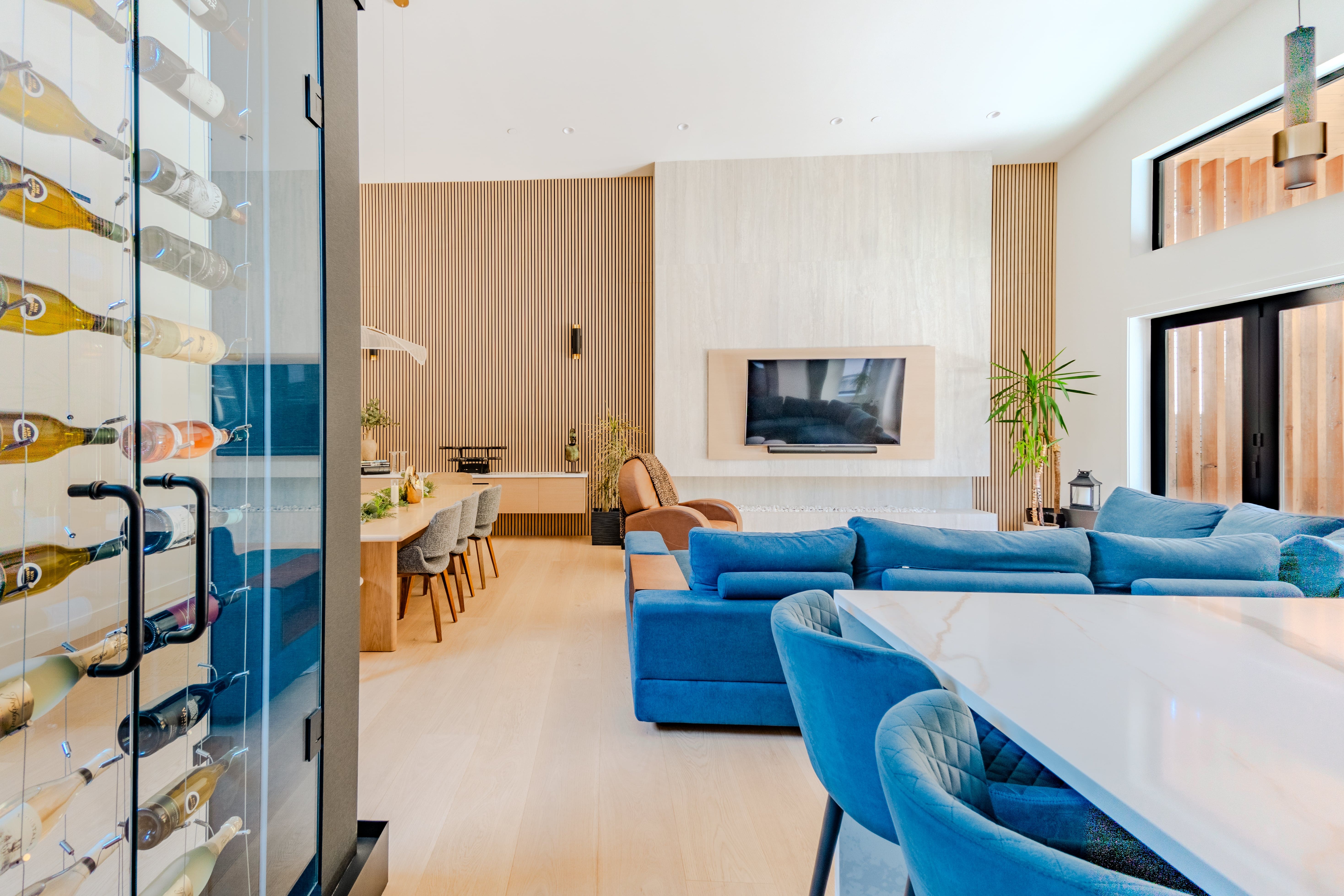 Open-plan living and dining real estate photography Vancouver — glass wine display wall, blue velvet sectional, marble dining table with velvet chairs, wood slat feature wall, and vaulted ceiling