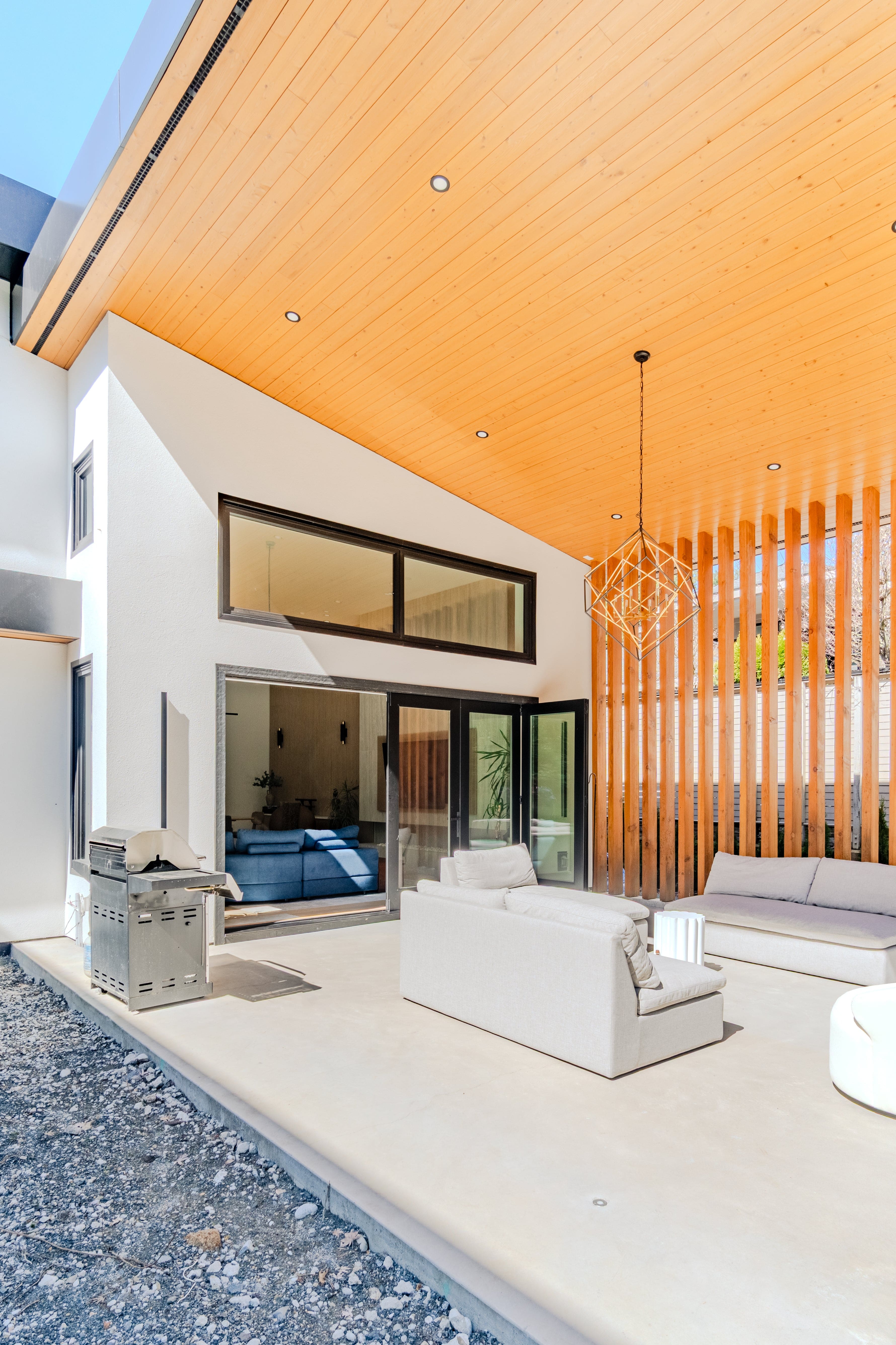 Covered outdoor patio real estate photography Vancouver — cedar tongue-and-groove ceiling, diamond geometric pendant, white lounge seating, glass sliding doors opening to interior, and vertical wood slat privacy screen