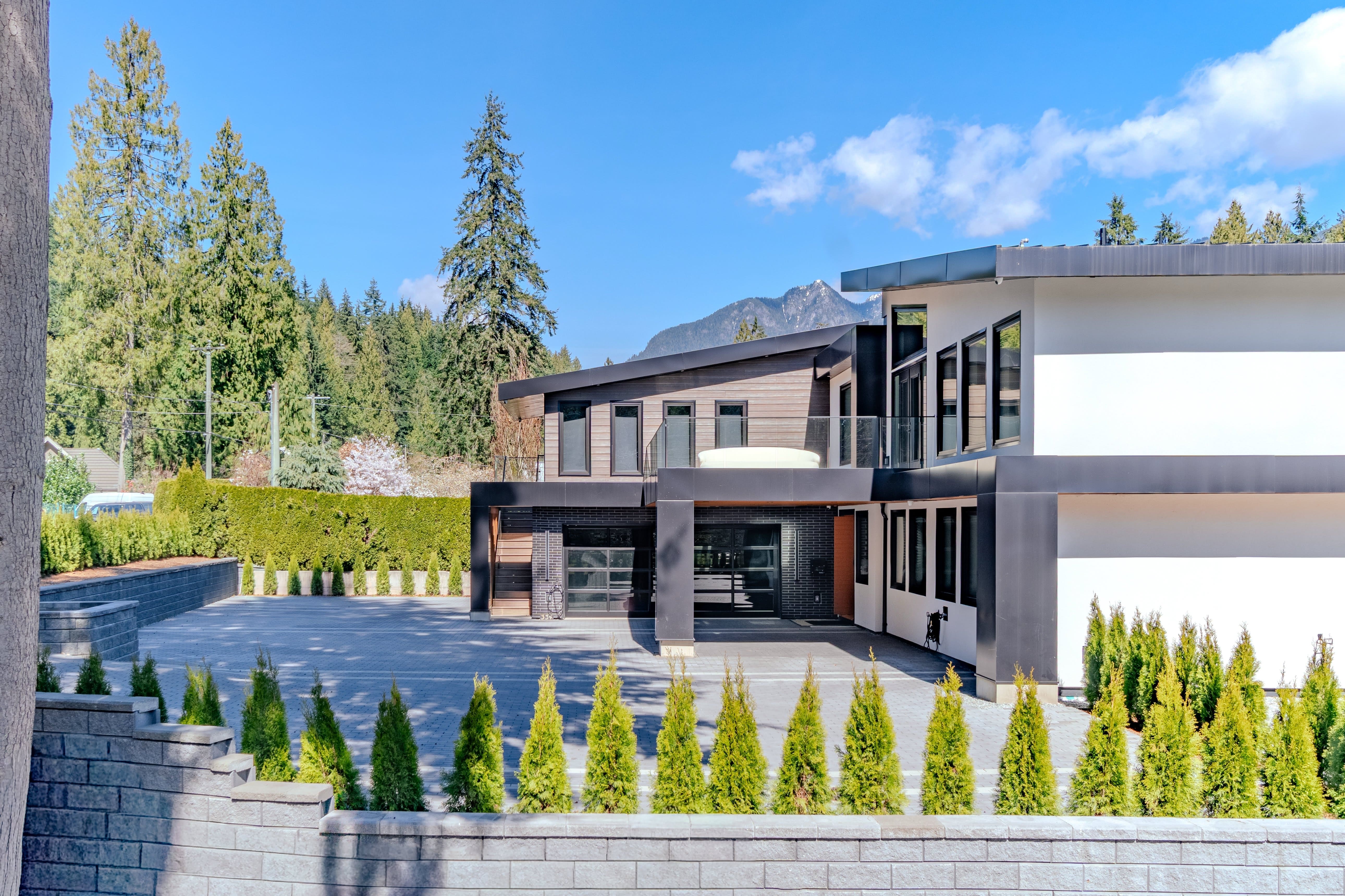 Modern luxury home exterior real estate photography Vancouver — dark charcoal and cedar facade, large glazing, paver driveway, columnar trees, cedar hedging, and snow-capped mountain backdrop
