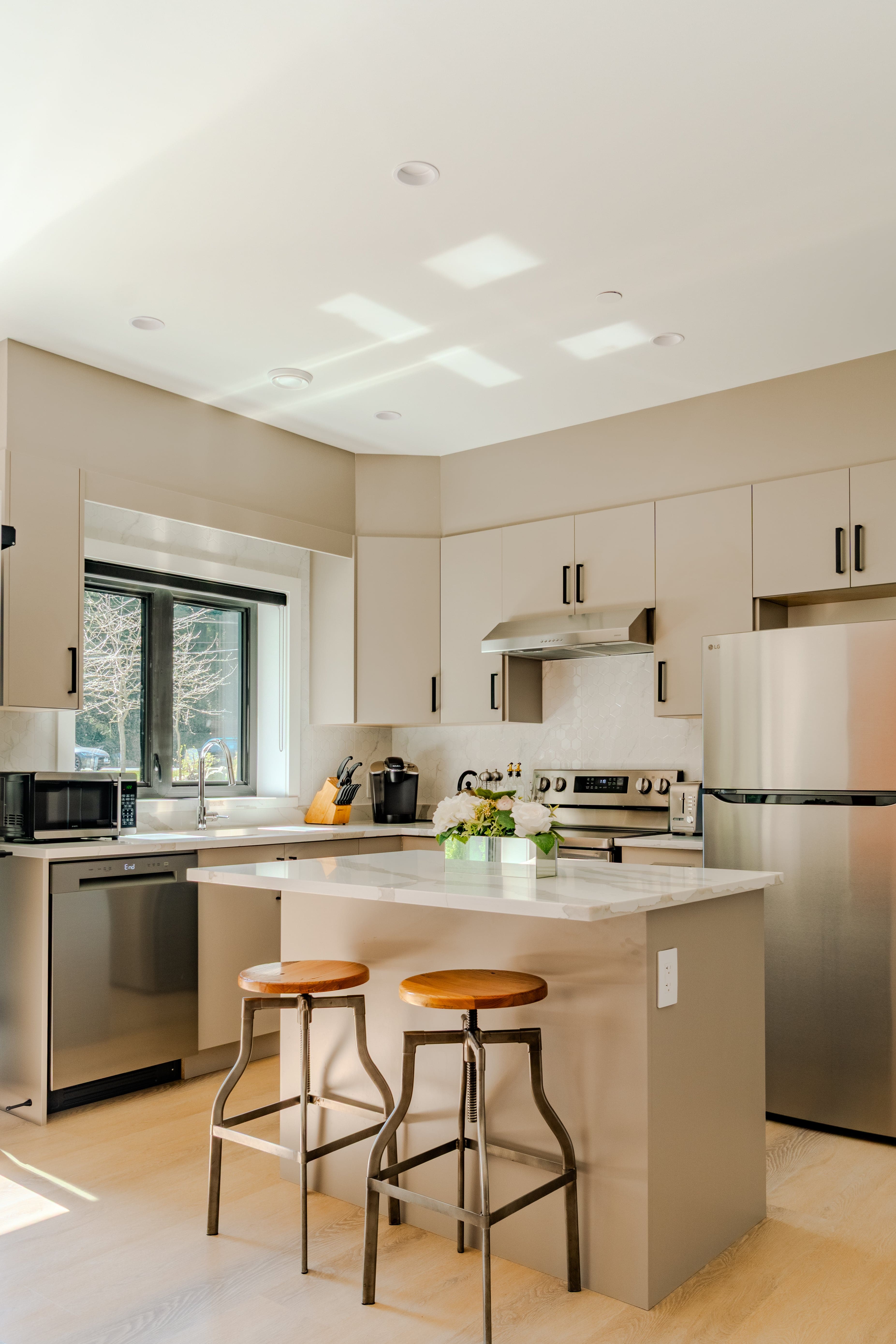 Kitchen real estate photography Vancouver — taupe flat-front cabinetry, quartz island with industrial wood and metal bar stools, stainless steel appliances, skylights, and garden window