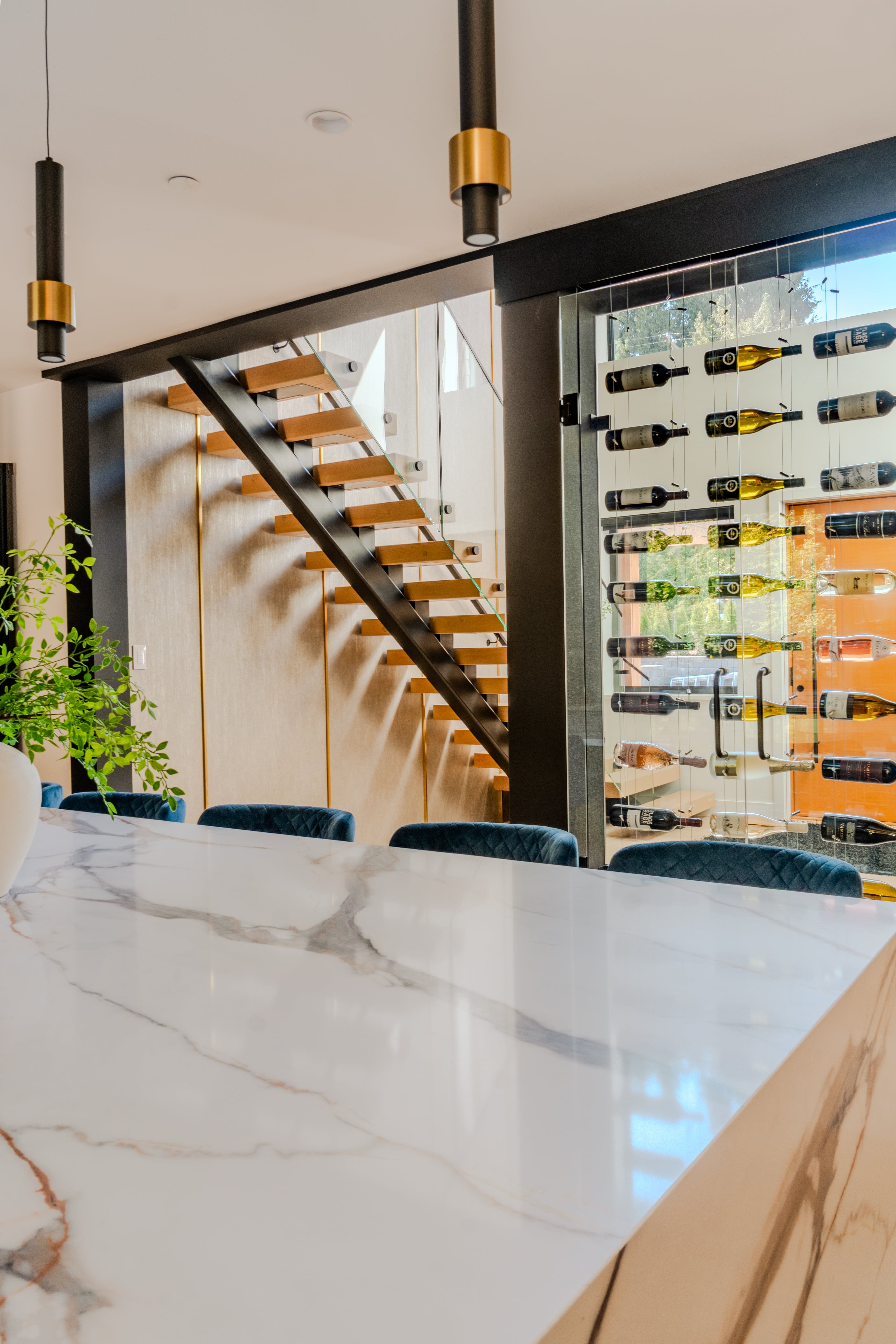 Dining area real estate photography Vancouver — white marble waterfall island, floor-to-ceiling glass wine display wall, floating staircase with brass cylinder pendants, and teal velvet dining chairs