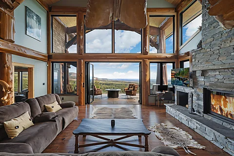 Open post-and-beam living room with a stone fireplace and forest views